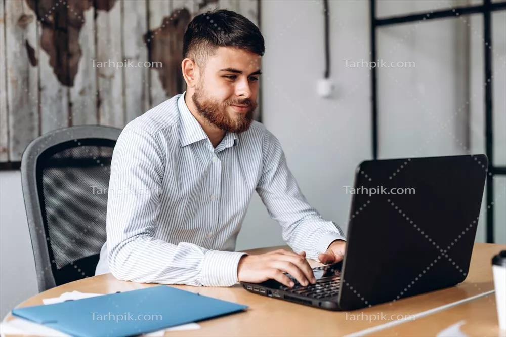 عکس مردی جدی با ریش در حال کار بر روی کامپیوتر در دفتر کار