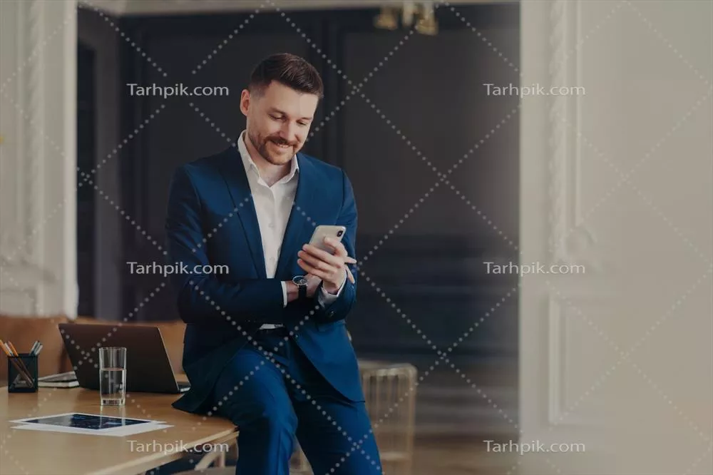 عکس مدیر خوشحال و خوش تیپ در حال بررسی موبایل خود در دفتر کار