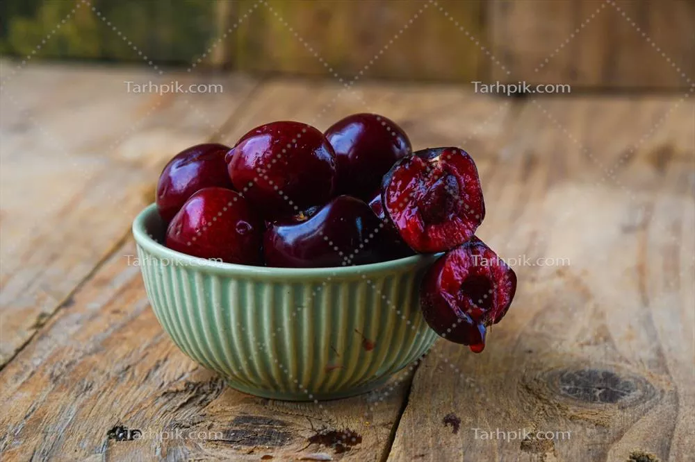 عکس چیدمان مینیمالیستی گیلاس های رسیده در کاسه سرامیکی سبز بر روی پس زمینه چوبی