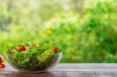 عکس سالاد تازه و رنگارنگ در کاسه چوبی بر روی میز با پس زمینه ای از برگ های سبز