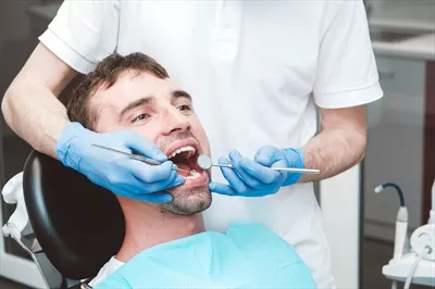 عکس دندانپزشک در حال معاینه دندان های یک بیمار مرد در صندلی دندانپزشکی