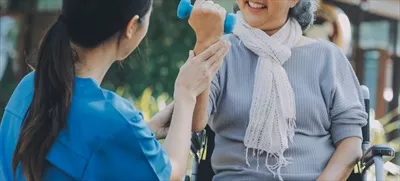 عکس پرستار جوان یا فیزیوتراپیست در حال کمک به یک زن سالخورده خوشحال برای انجام تمرینات ورزشی در خانه