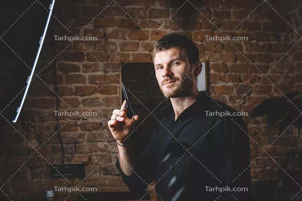 عکس آرایشگر مرد با قیچی تیز در برابر دیوار آجری ایستاده است