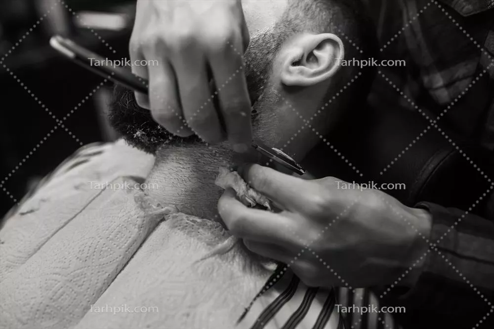 عکس مرد جوان با ریش در حال اصلاح توسط آرایشگر در آرایشگاه