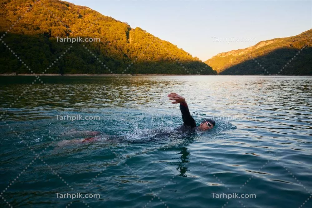 عکس ورزشکار تریاتلون در حال شنا در دریاچه در طلوع آفتاب با لباس مخصوص