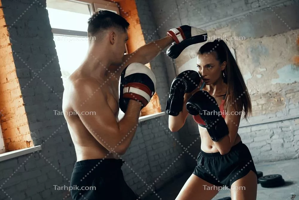 عکس زنی جوان و متمرکز در حال تمرین بوکس با مربی شخصی در باشگاه