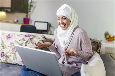 عکس زنی جوان و مسلمان در حال برقراری تماس ویدیویی با لپ تاپ در خانه