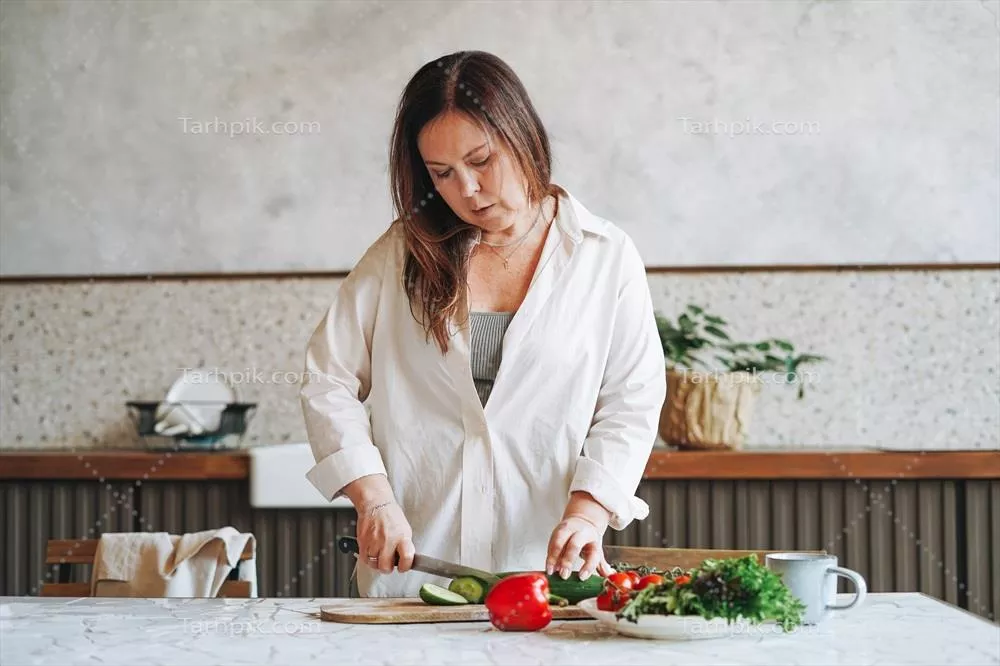 عکس زنی میانسال با موهای قهوه ای در حال تهیه سالاد سبزیجات تازه در آشپزخانه