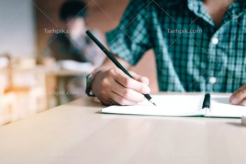 عکس نزدیک نمایی از دست یک دانش آموز در حال نوشتن بر روی کاغذ با مداد در کلاس درس