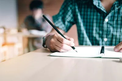 عکس نزدیک نمایی از دست یک دانش آموز در حال نوشتن بر روی کاغذ با مداد در کلاس درس