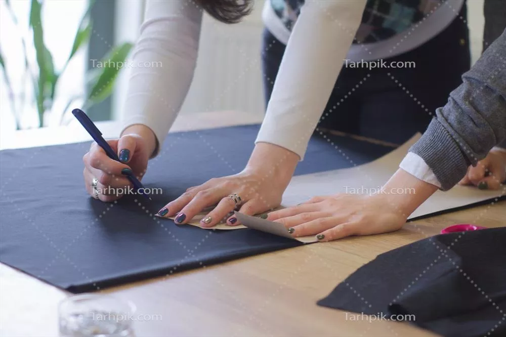 عکس زنی خیاط در حال کار بر روی میز، مشغول دوخت و دوز لباس در استودیو