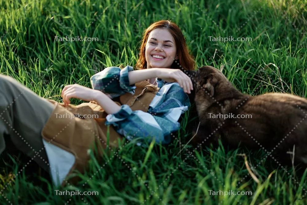 زنی که در پارک در هنگام غروب تابستانی بر روی چمن دراز کشیده و با سگ خود cuddle می‌کند