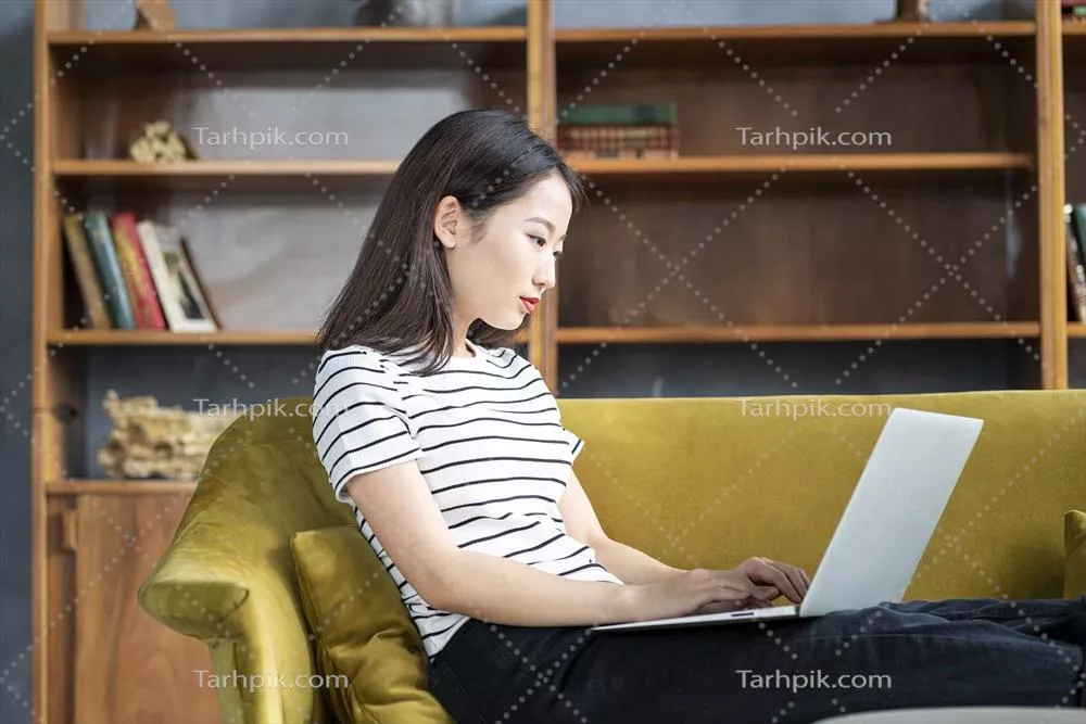 زنی ژاپنی زیبا در حال لبخند زدن و تایپ کردن بر روی کامپیوتر در هتلی بر روی کاناپه