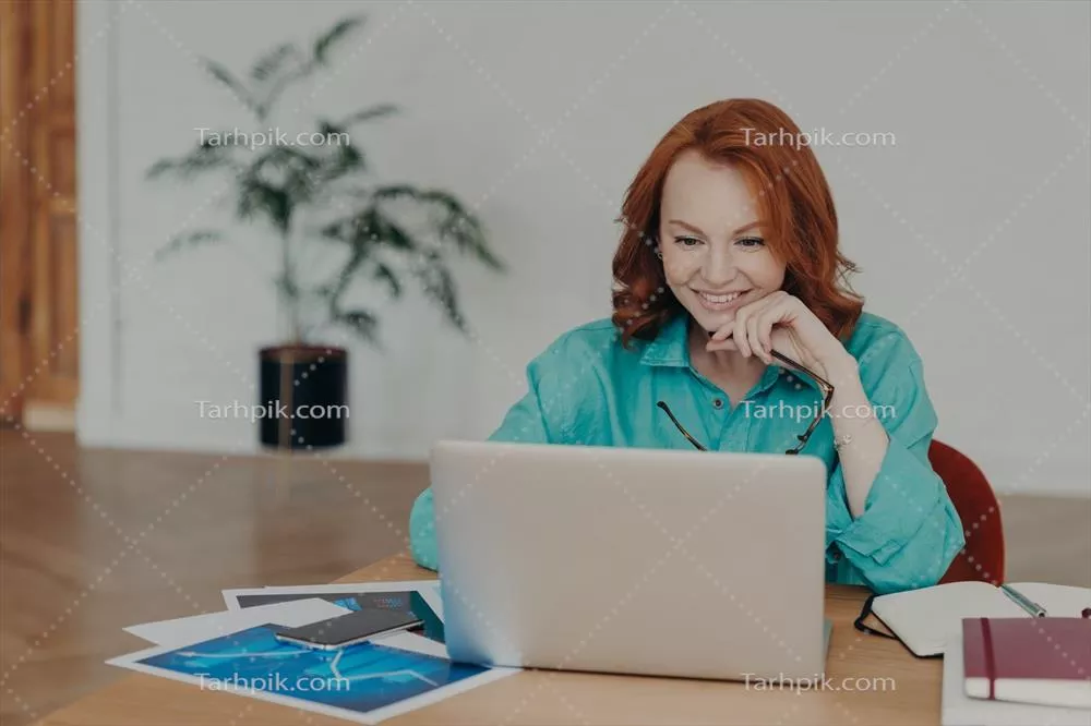 زنی جوان با موهای قرمز، در حال کار یا تحصیل با لپ‌تاپ مدرن و دریافت مشاوره آنلاین