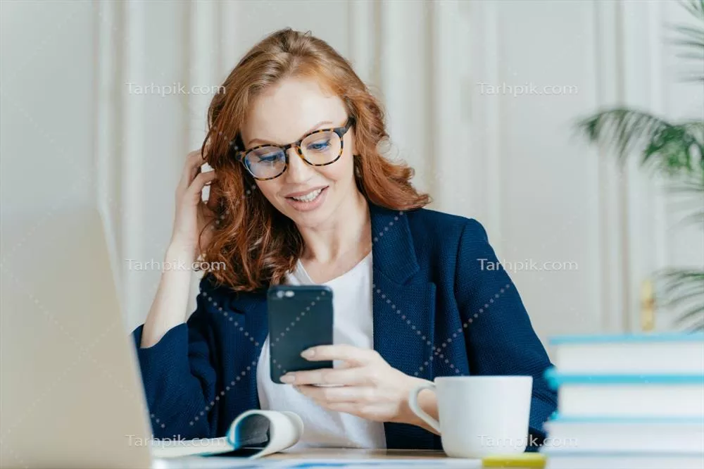 یک لنسری زن با موهای فر قرمز و عینک در حال استفاده از تلفن هوشمند برای ارتباط آنلاین