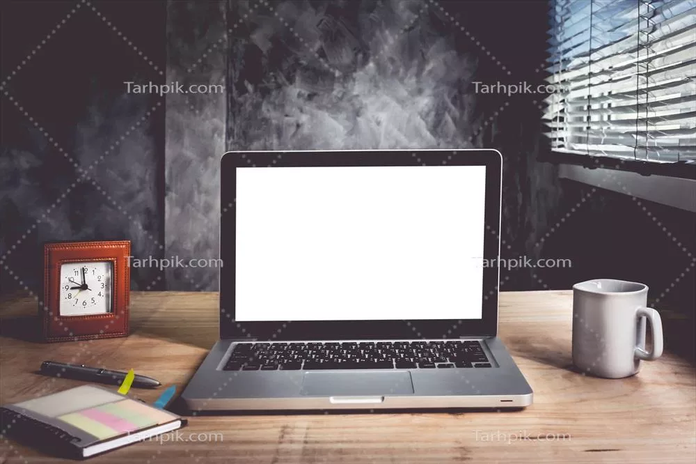 نمای نزدیک از لپتاپ با صفحه سفید و فنجان قهوه و دفتری بر روی میز چوبی در پسزمینه دیوار فرسوده