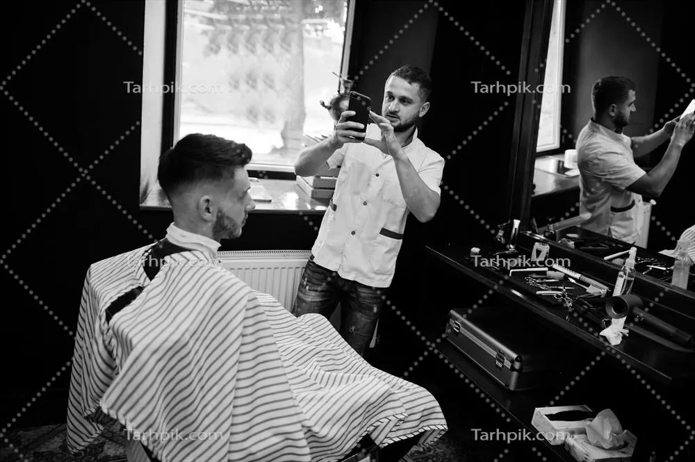 مرد جوان با ریش در حال کوتاهی مو توسط آرایشگر در آرایشگاه در حال صحبت کردن در تلفن
