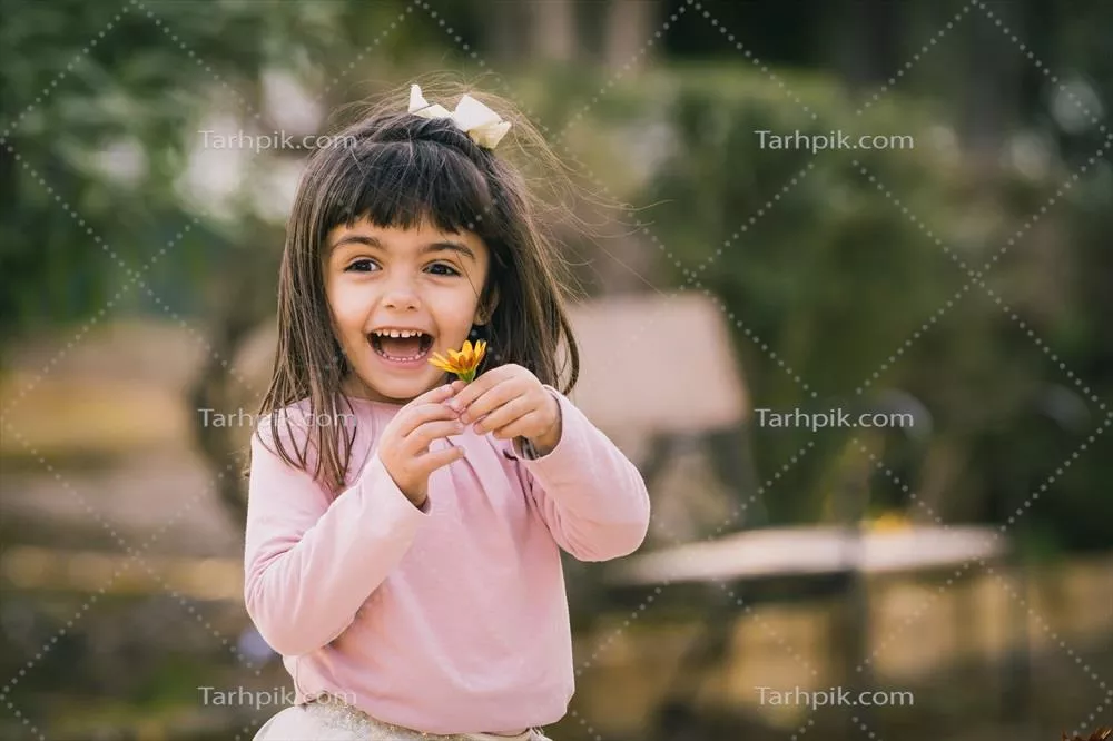 دختری کوچک در پارک در حال گرفتن یک گل با لبخند شاداب و خوشحال