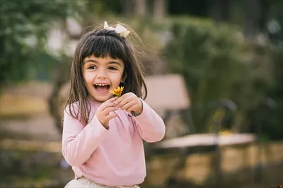 دختری کوچک در پارک در حال گرفتن یک گل با لبخند شاداب و خوشحال
