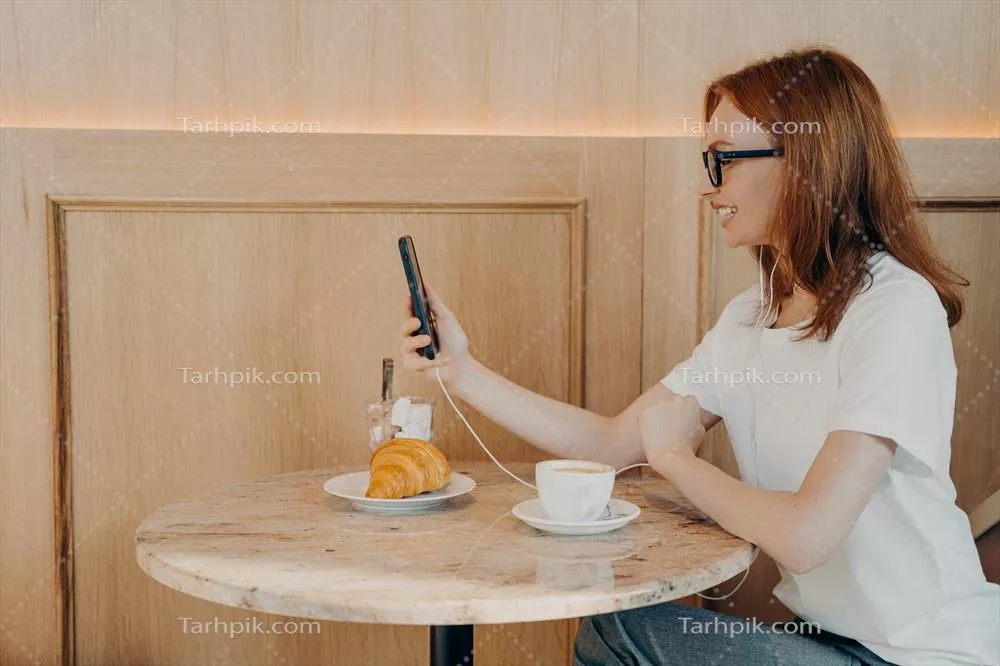 زن جوان قرمز مو و شاد که هنگام نشستن در کافه از اینترنت رومینگ استفاده می کند