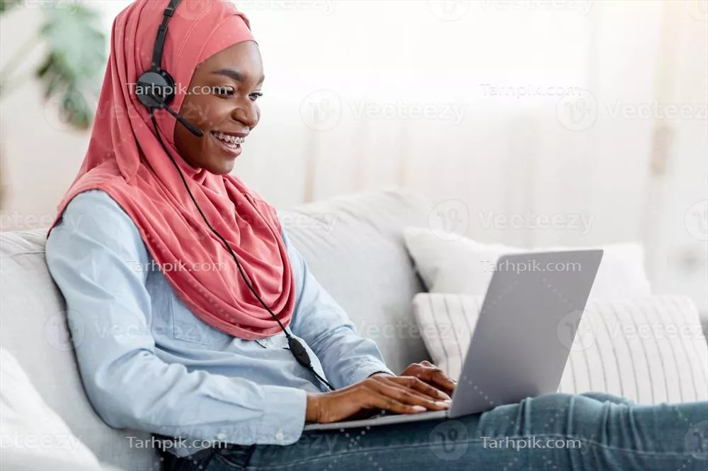 آموزش آنلاین: زن مسلمان خوشحال آفریقایی با حجاب و هدفون در حال مطالعه در خانه