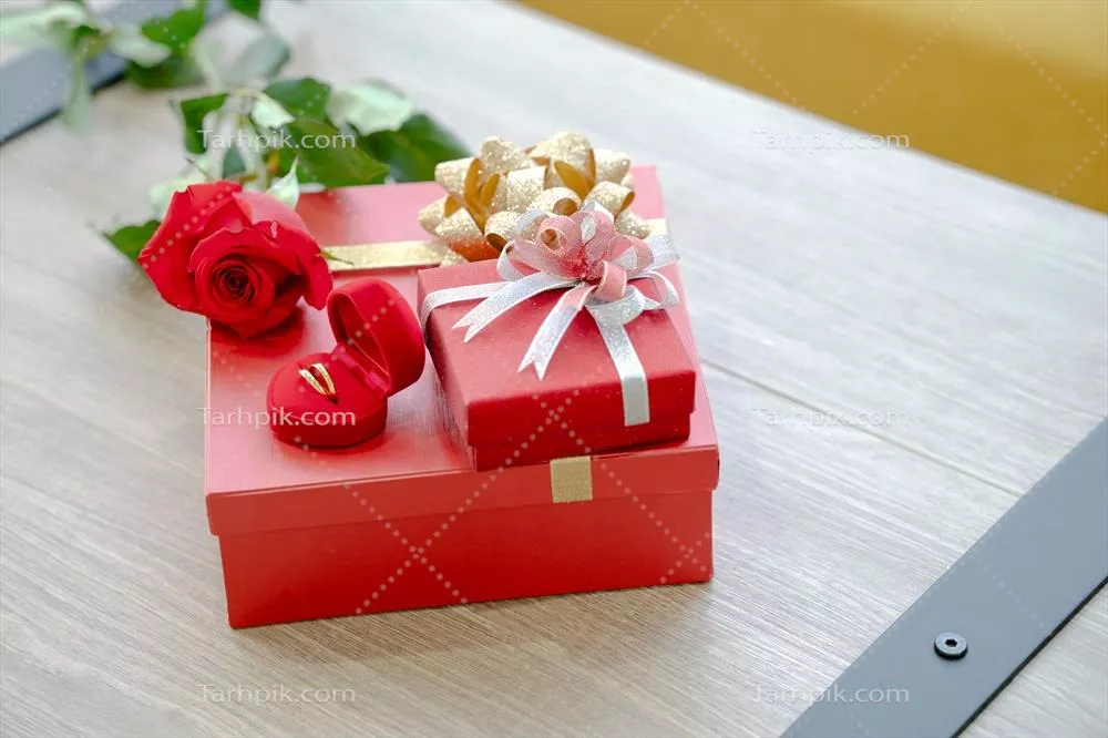 جعبه هدیه با روبان قرمز و حلقه گل طلایی و گل رز روی میز چوبی