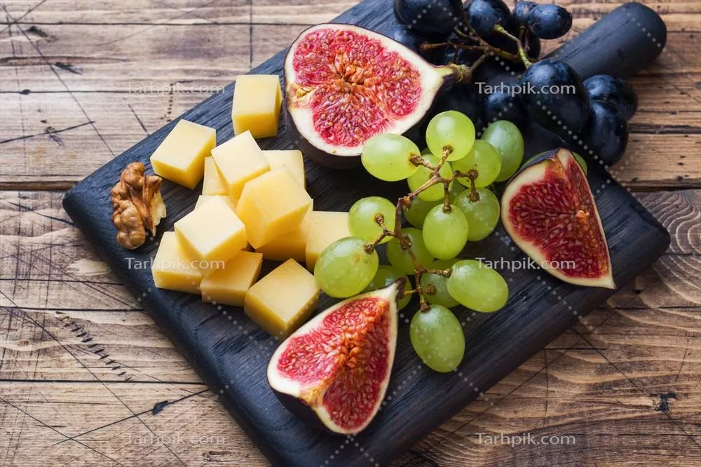 مکعبهای پنیر تازه، میوههای تازه، انجیر، انگور، عسل و گردو بر روی تخته برش چوبی