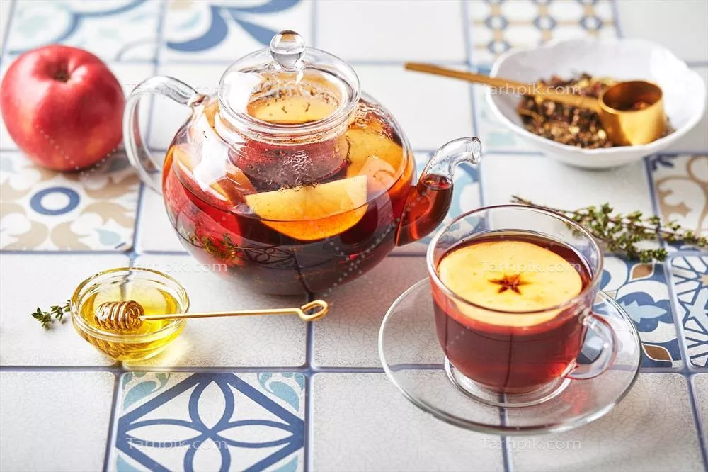 چای میوهای با سیب و آویشن در قوری شیشهای و فنجان بر روی میزی از کاشیهای رنگی