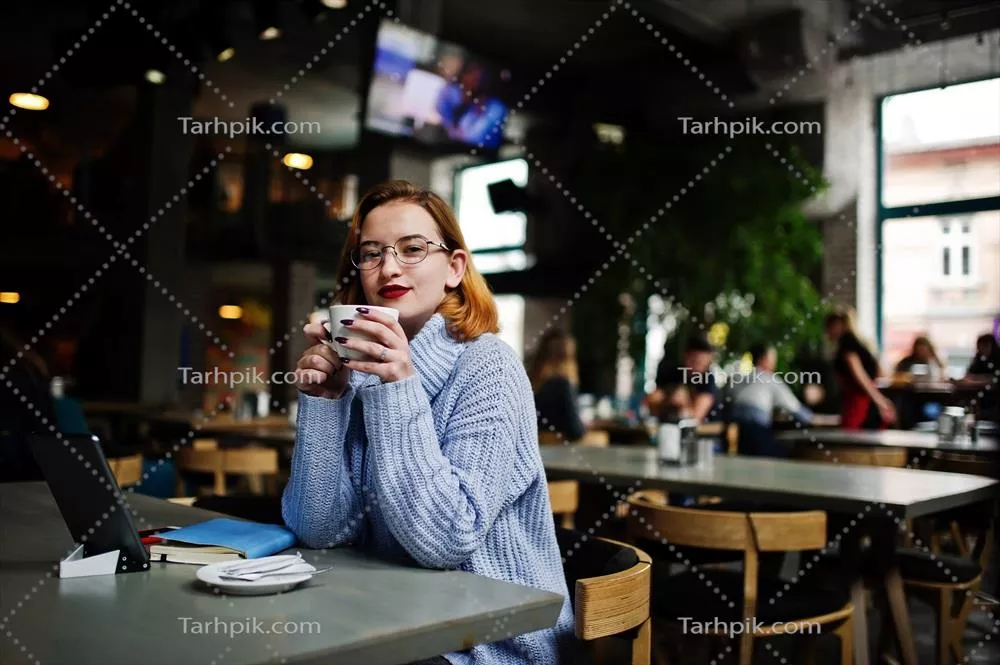 عکس زنی جوان و خوشحال با موهای قرمز و عینک که در کافه مشغول کار است