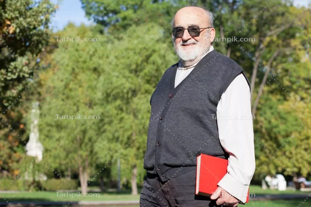 عکس مرد سالخورده با عینک آفتابی در پارک در حال نگه داشتن کتاب قرمز