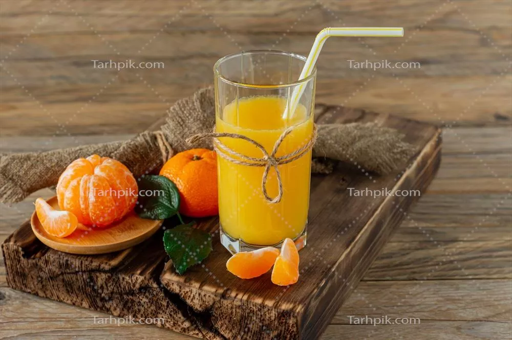 عکس نگاهی به یک لیوان آب پرتقال ترد همراه با پرتقال های رسیده بر روی تخته چوبی