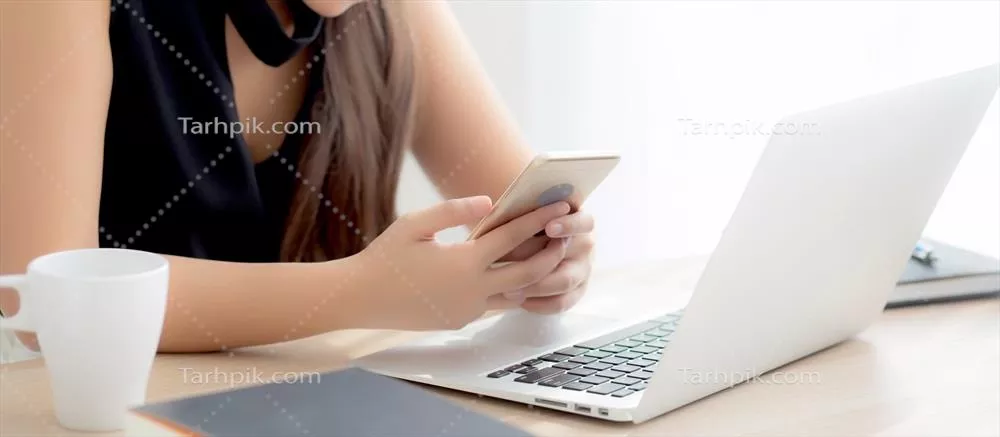 عکس زنی جوان و زیبا از آسیای شرقی که به لپ‌تاپ مشغول به کار است و از تلفن هوشمند خود استفاده می‌کند